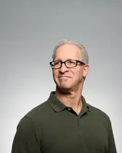 Photo of a smiling older man with short gray hair and rectangular glasses against a plain grey backdrop