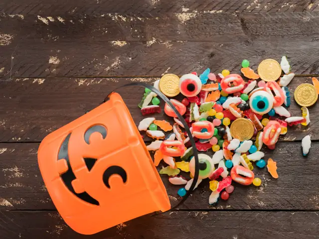An orange jack-o'-lantern bucket tipped over on a wooden table, spilling gummy eyeballs, sweets and chocolate coins