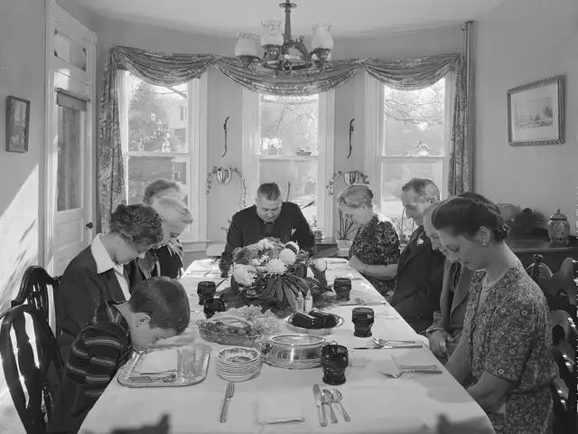 A 1942 black-and-white photograph: a family of eight seated with bowed heads at a formally laid Thanksgiving table with white cloth and centrepiece