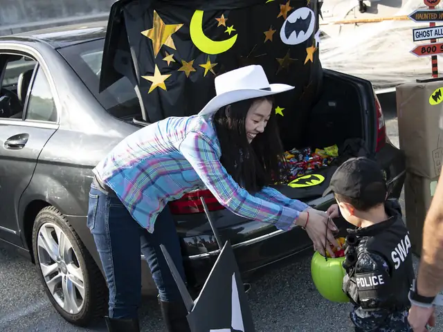 A car boot decorated with paper stars and a moon at a trunk-or-treat event, a woman in a cowboy hat handing candy to a small boy in a police costume