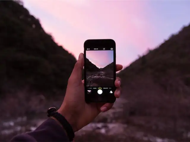 A hand holding up a phone to photograph a pink evening sky between two hills