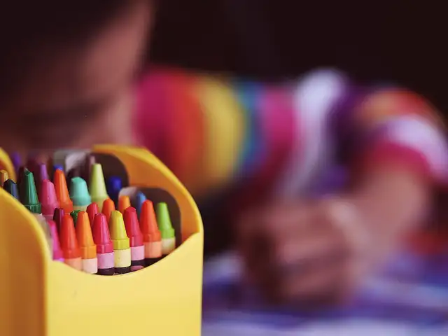 A yellow box of crayons in sharp focus in the foreground, and beyond it a child in a rainbow-striped shirt leaning over paper, coloring, blurred