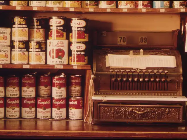 Old brass cash register with price flags reading 70 and 9 cents, canned goods on wooden shelves of a mid-century shop