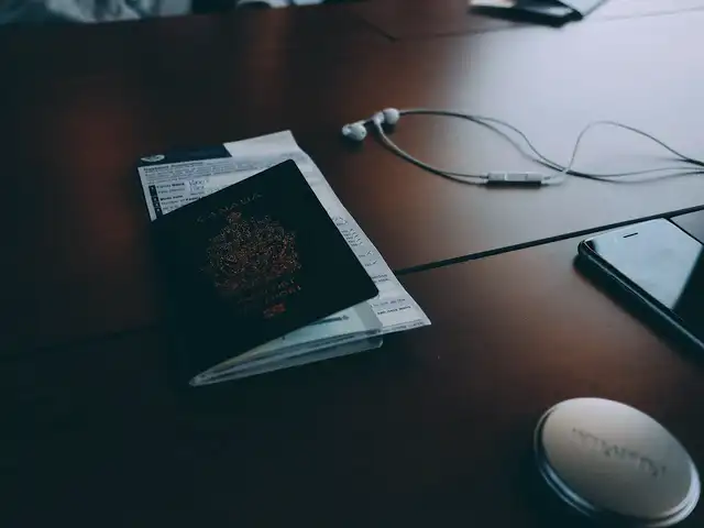 A dark wooden desk with a closed passport lying on a paper form, beside earbuds, a phone and a small round tin
