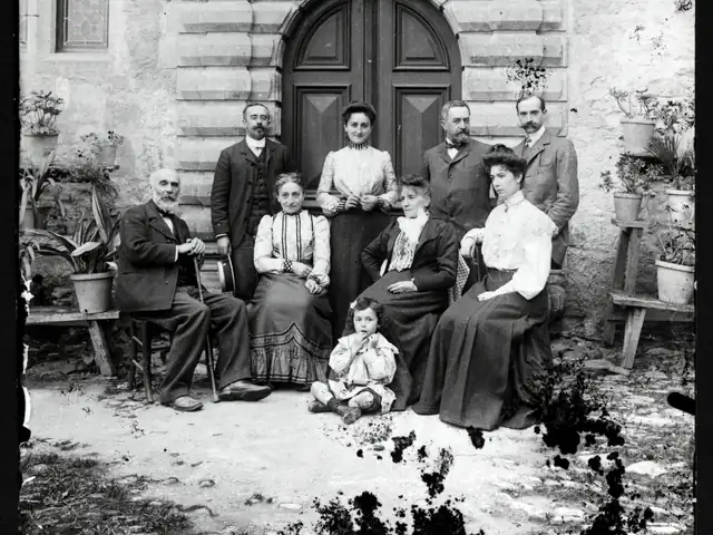 Family of nine across three generations, from a bearded grandfather to a toddler seated on the ground, posed in 1905 before a carved stone doorway — the old print faded, with dark blotches eating at its edges