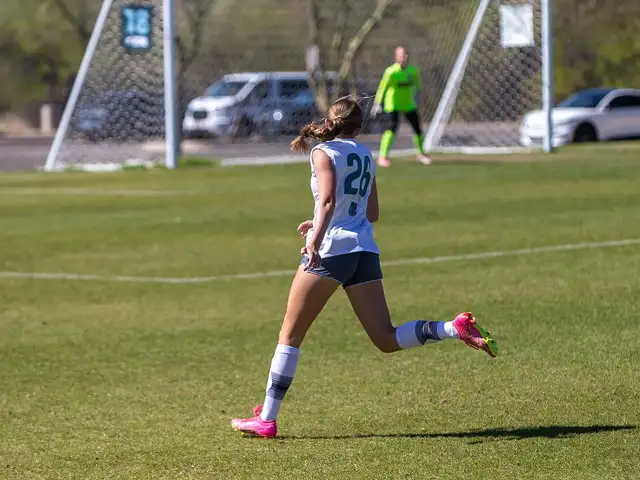A youth player in a white training bib with the number 26 running toward a goal, photographed from behind on a sunny grass field